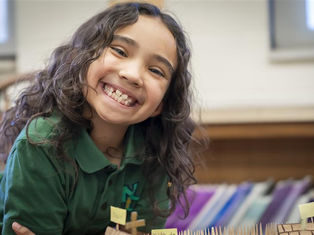 Smiling child with curly hair in a green shirt leans over a table with labeled mini flags. Classroom setting with colorful folders in background.