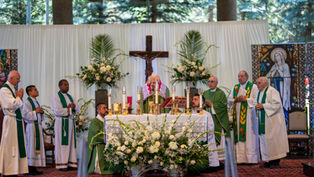 Clergy in green vestments conduct a Catholic Mass at an altar with candles and flowers, under a crucifix.