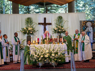 Clergy in green vestments conduct a Catholic Mass at an altar with candles and flowers, under a crucifix.