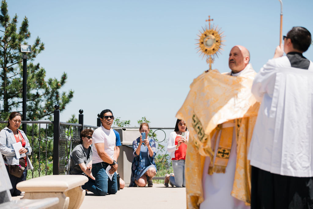 Outdoor Catholic prayer service with priest holding a monstrance while people kneel and take photos on a sunny terrace.