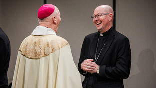 Two clergy members, one in a cream robe with a pink cap, the other in black attire, share a joyful conversation in a neutral setting.