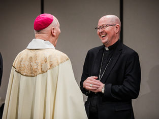 Two clergy members, one in a cream robe with a pink cap, the other in black attire, share a joyful conversation in a neutral setting.