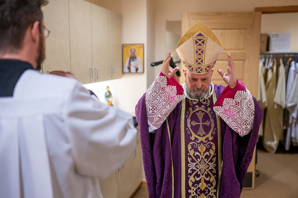 A bishop in ornate purple vestments and mitre gestures solemnly in a room, facing a priest in white. Religious decor in the background.