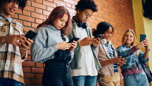 Five teens with phones lean against a brick wall. They are focused on screens, some smiling. Casual attire, headphones visible.