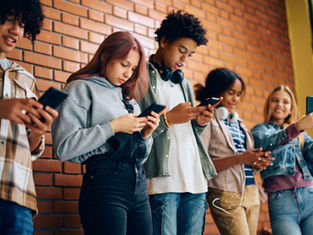 Five teens with phones lean against a brick wall. They are focused on screens, some smiling. Casual attire, headphones visible.