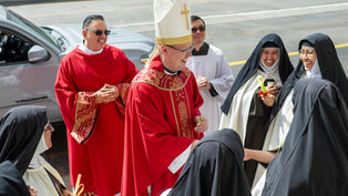 Bishop in red vestments and mitre smiles with nuns and clergy on a sunny street, exchanging palm crosses.