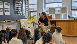 Teacher reads a colorful book to attentive children in a classroom. Chalkboard, posters, and text on easel in the background. Cozy setting.