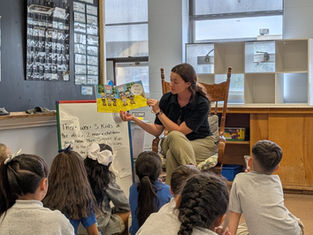 Teacher reads a colorful book to attentive children in a classroom. Chalkboard, posters, and text on easel in the background. Cozy setting.