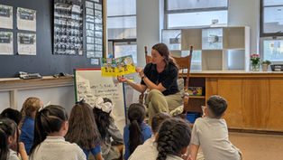 Teacher reads a colorful book to attentive children in a classroom. Chalkboard, posters, and text on easel in the background. Cozy setting.