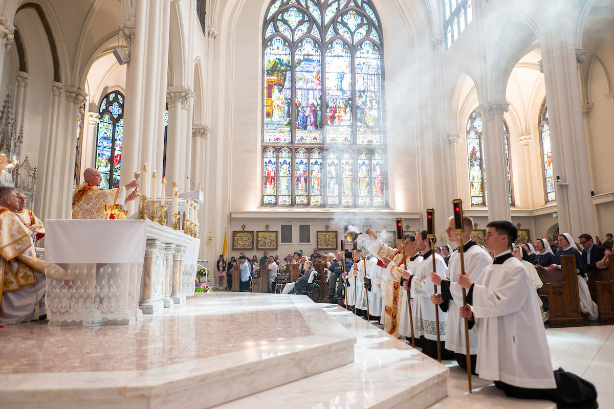 Clergy and altar servers kneel near the altar during Mass as incense rises in the background.