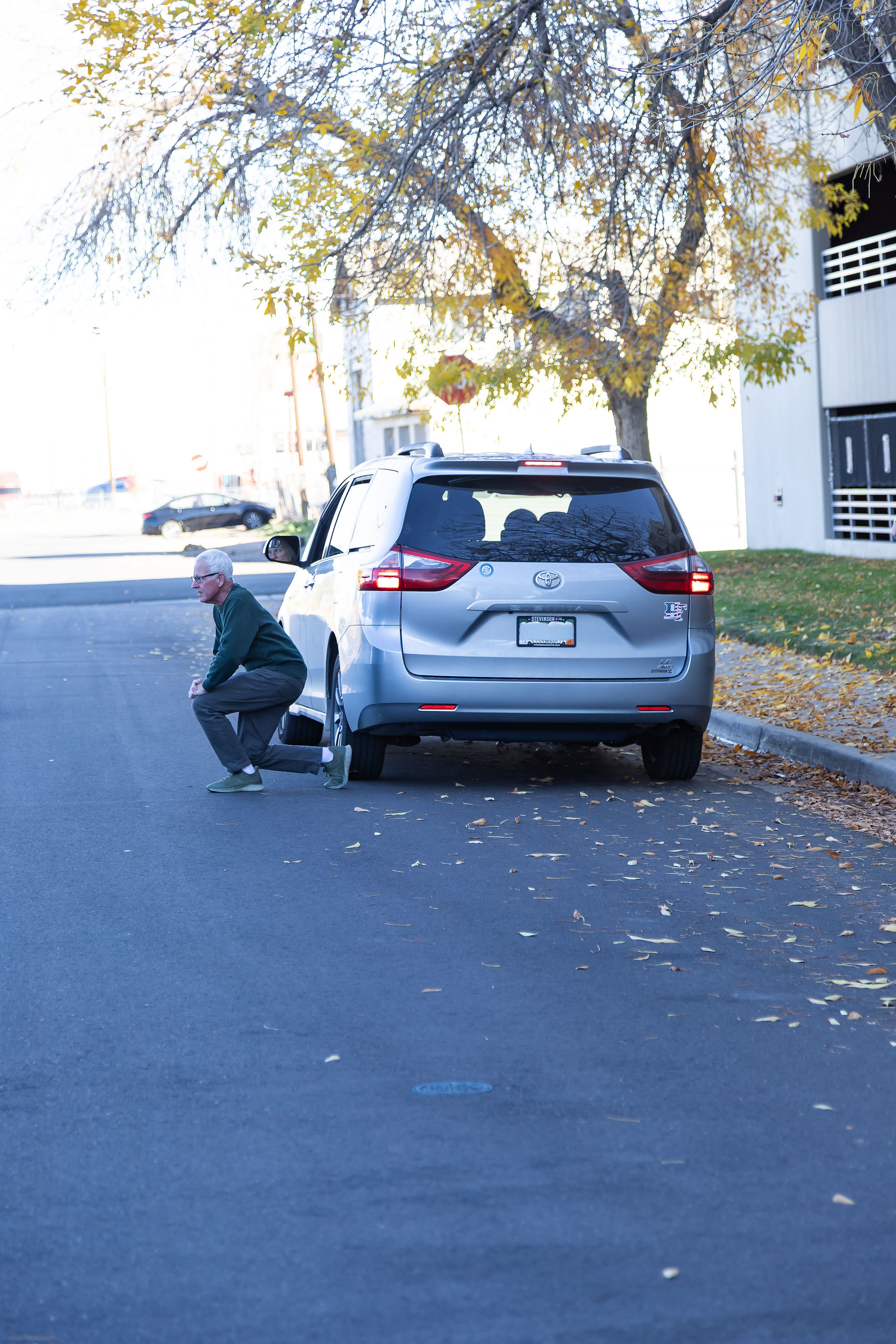 Elderly man kneeling beside a parked silver Toyota on a street lined with autumn leaves, wearing green shoes. Trees with yellowing leaves in background.