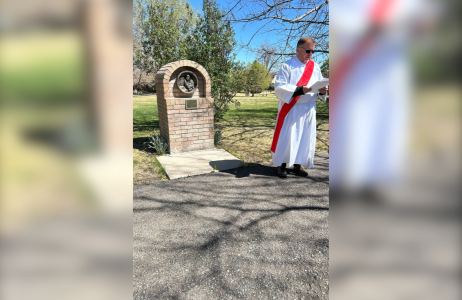 A person in a white robe with a red sash reads a document near a brick monument outdoors on a sunny day, with trees and grass in the background.