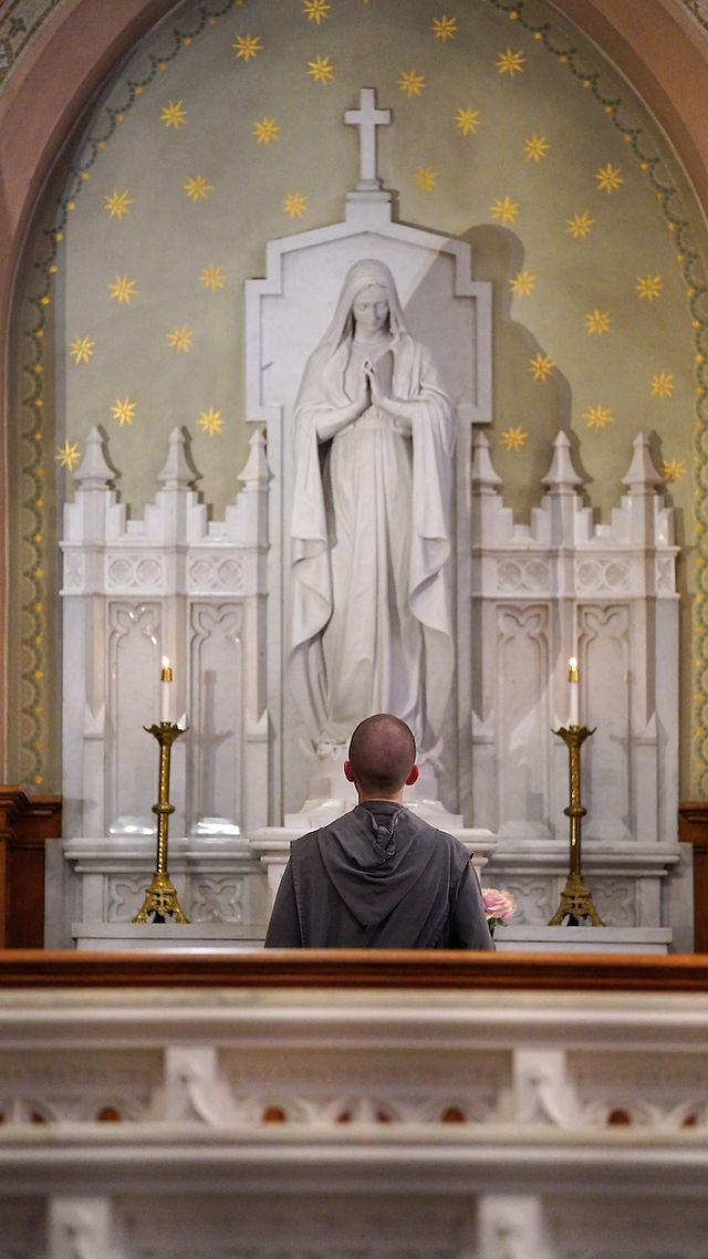A person in a robe stands before a white statue of a praying figure in a church. Stars adorn the wall; candles flank the altar. Peaceful mood.
