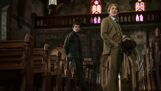 Two men stand in a dimly lit church with stained glass windows. One in a suit holds a hat, looking serious. Wooden pews line the aisle.