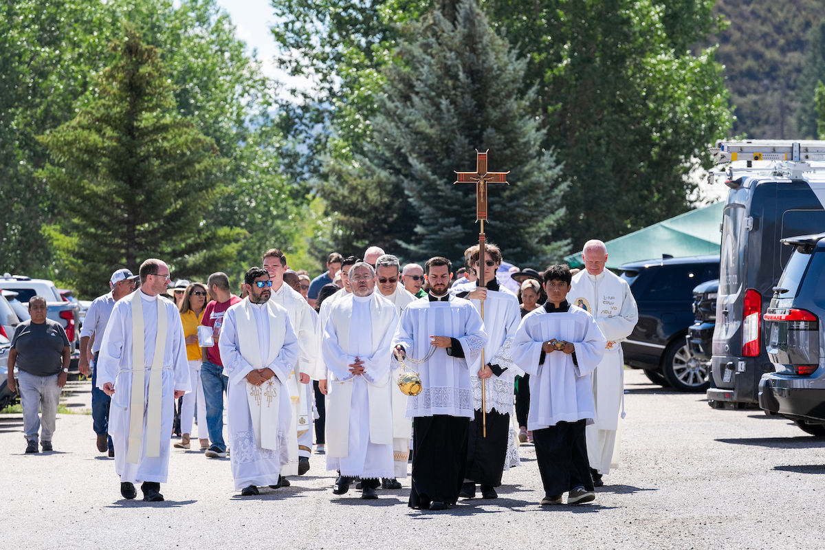 Clergy in white robes lead an outdoor religious procession with a cross and incense, walking along a tree-lined street.