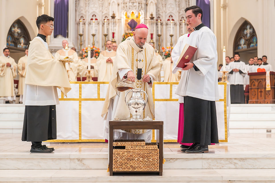 Clergy in white robes perform a ritual pouring liquid from glass jugs at an ornate altar with candles and flower arrangements in a church.