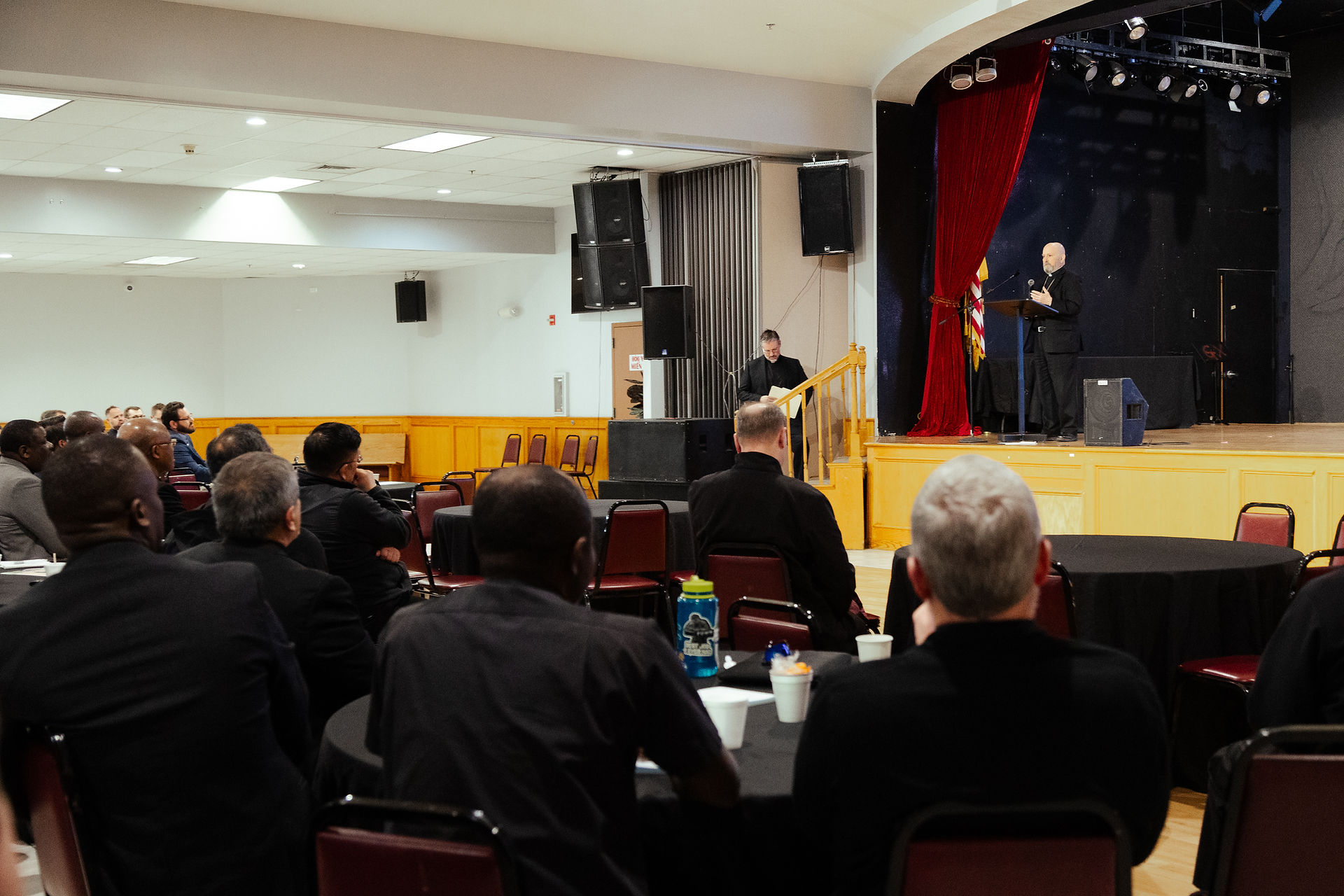 A speaker addresses a seated audience in a hall with a stage, red curtains, and black tables. People appear attentive and engaged.