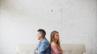 Two people sit back-to-back on a beige sofa, arms crossed, against a white brick wall. They appear upset or distant.