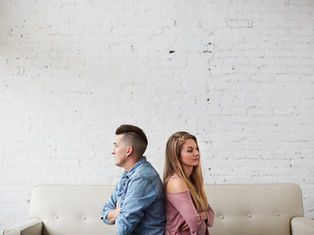 Two people sit back-to-back on a beige sofa, arms crossed, against a white brick wall. They appear upset or distant.