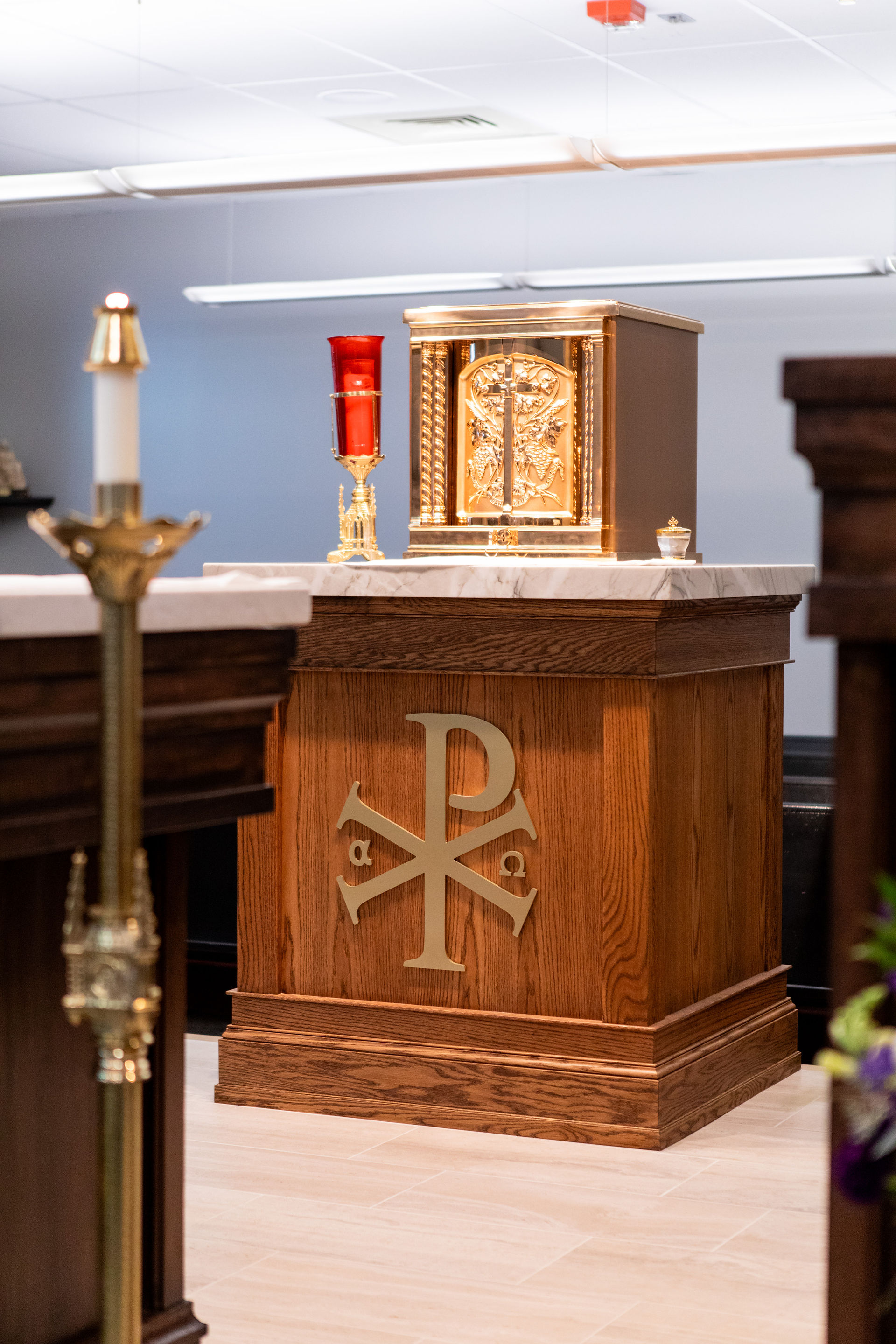 Wooden altar with a golden tabernacle and red candle in a church. Chi Rho symbol on altar front. Calm, reverent setting.