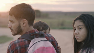 A man with a backpack carries a child, standing beside a woman. All wear plaid shirts in a serene rural landscape at sunset, looking thoughtful.