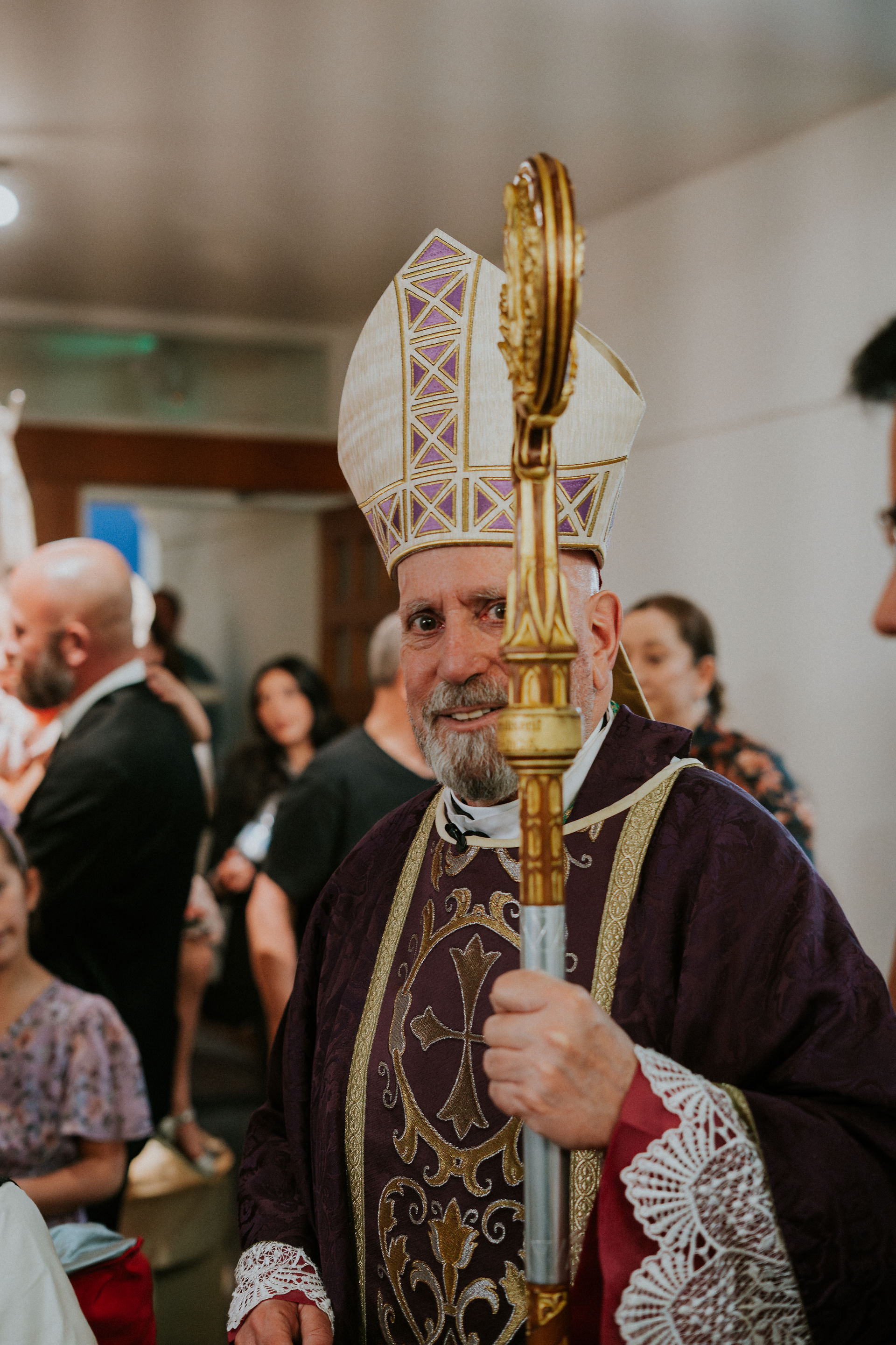 Bishop in purple and gold vestments with ornate miter, holding a crozier, surrounded by people in a church setting, smiling warmly.
