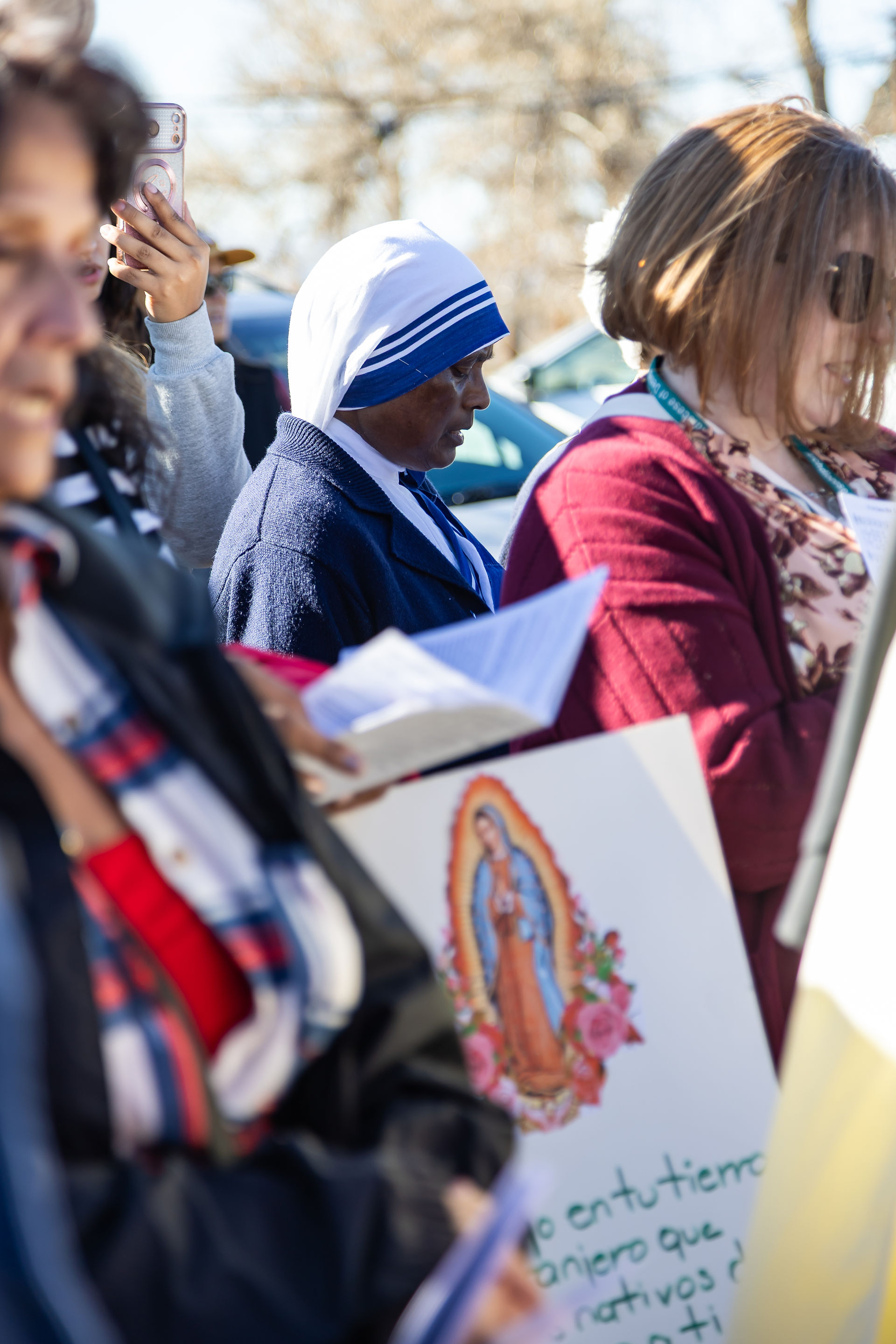 People gathered outdoors, one in a blue and white habit, holding papers. A sign with a religious image and text visible. Sunny day.