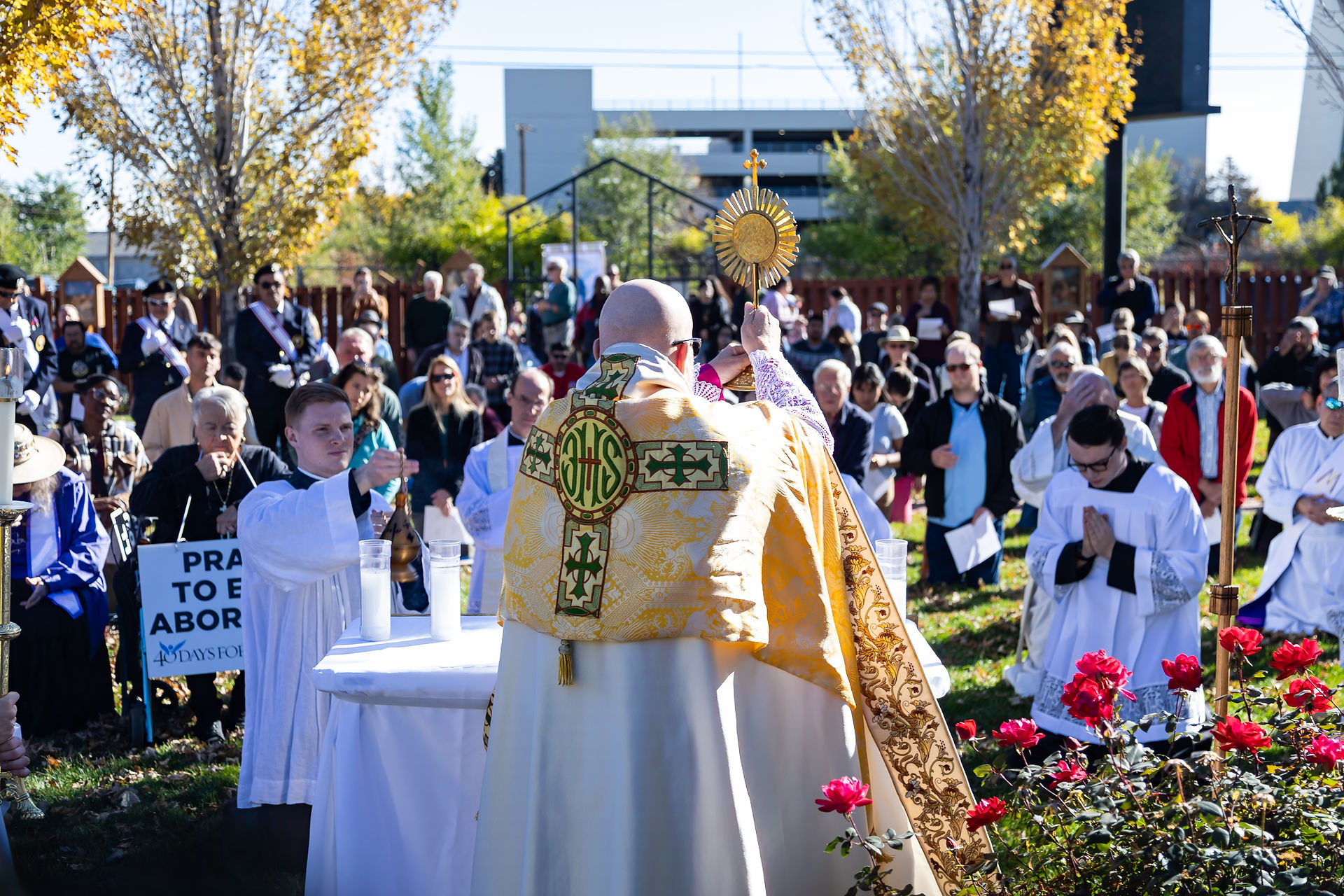Archbishop in ornate vestments holds a monstrance. Crowd gathers outdoors with flowers and trees. "PRAY TO END ABORTION" sign visible.