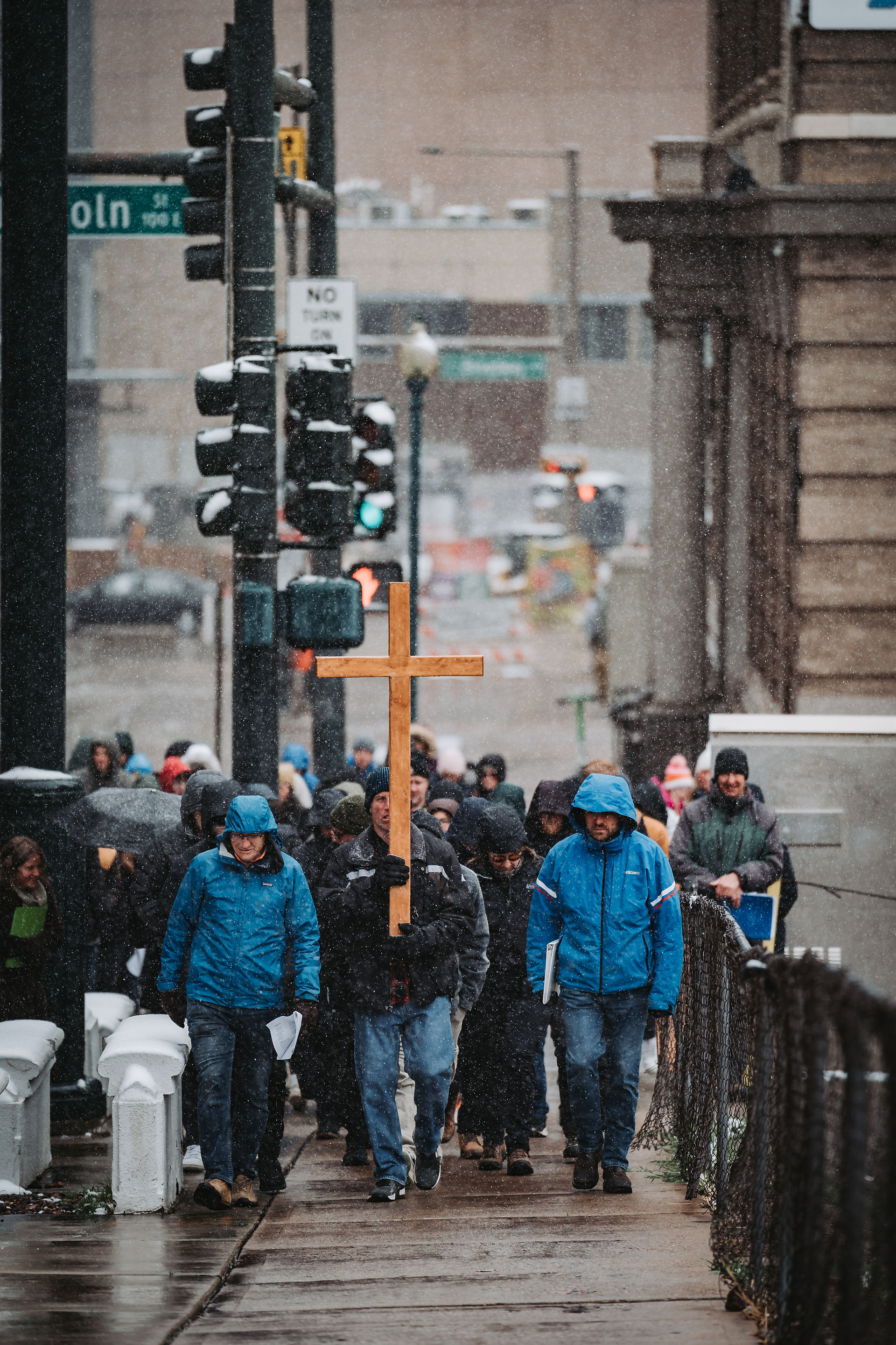People in winter coats walk on a snowy street, one carrying a wooden cross. Street signs and buildings visible in the background.