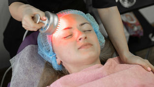 Woman receives facial treatment with a red light device. She is wearing a blue hair cap and pink towel in a clinical setting. Relaxed mood.