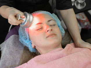 Woman receives facial treatment with a red light device. She is wearing a blue hair cap and pink towel in a clinical setting. Relaxed mood.