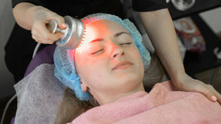 Woman receives facial treatment with a red light device. She is wearing a blue hair cap and pink towel in a clinical setting. Relaxed mood.