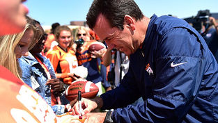Man in a blue jacket signs a football for a fan in a crowd wearing orange. Daylight setting with a clear sky; cheerful atmosphere.