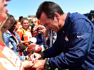 Man in a blue jacket signs a football for a fan in a crowd wearing orange. Daylight setting with a clear sky; cheerful atmosphere.