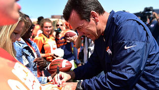 Man in a blue jacket signs a football for a fan in a crowd wearing orange. Daylight setting with a clear sky; cheerful atmosphere.