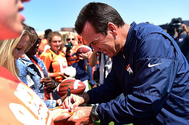 Man in a blue jacket signs a football for a fan in a crowd wearing orange. Daylight setting with a clear sky; cheerful atmosphere.