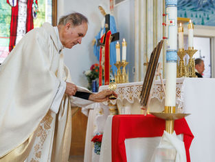 Priest in a white robe places an item on an altar with candles and religious icons. Stained glass window in the background creates a solemn mood.