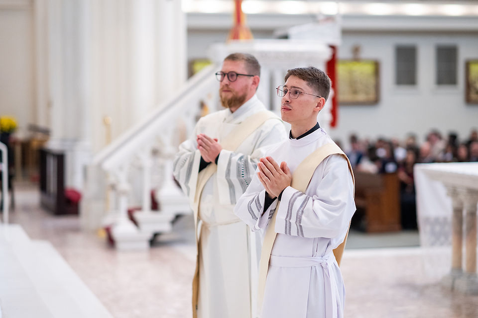 Two clergy in white robes stand with hands clasped in prayer in a church. The background shows pews and soft lighting. Quiet atmosphere.