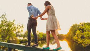 A couple balances on a wooden rail, holding hands. They're surrounded by trees and a sunset reflection on water, creating a serene mood.