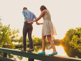 A couple balances on a wooden rail, holding hands. They're surrounded by trees and a sunset reflection on water, creating a serene mood.