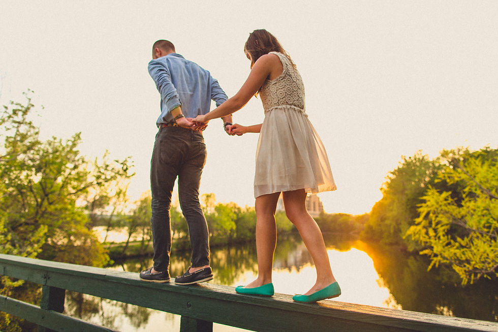 A couple balances on a wooden rail, holding hands. They're surrounded by trees and a sunset reflection on water, creating a serene mood.