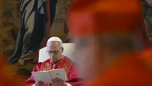 A man in red and white religious attire reads a booklet in a church with a mural background. Another figure in red appears blurred in the foreground.