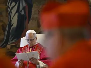 A man in red and white religious attire reads a booklet in a church with a mural background. Another figure in red appears blurred in the foreground.