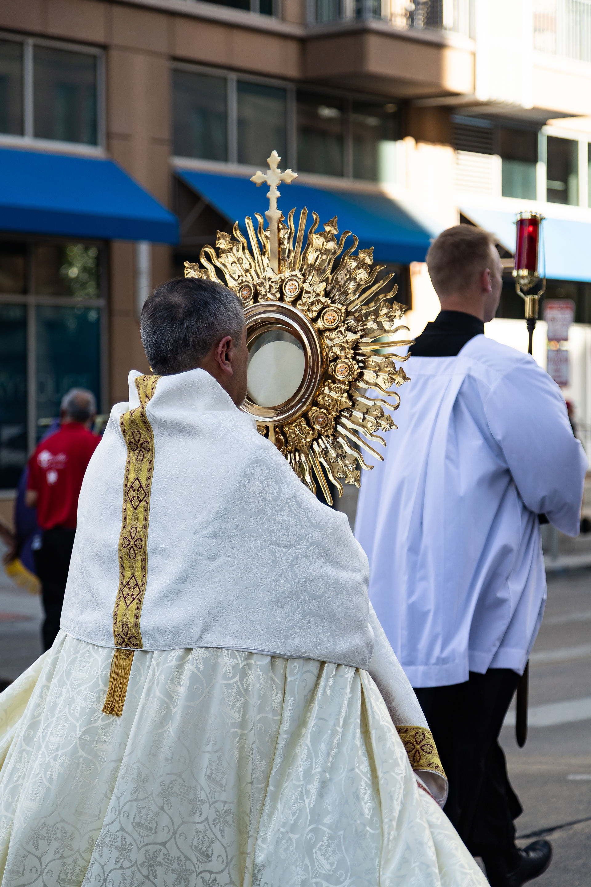 Catholic procession on a city street, with a priest carrying a golden monstrance and another server beside him in white robes.
