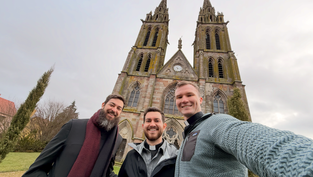 Three men smile in front of a gothic-style church with two tall towers. Overcast sky and bare trees in the background.