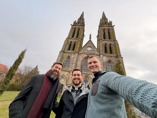Three men smile in front of a gothic-style church with two tall towers. Overcast sky and bare trees in the background.