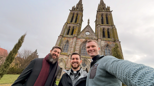 Three men smile in front of a gothic-style church with two tall towers. Overcast sky and bare trees in the background.