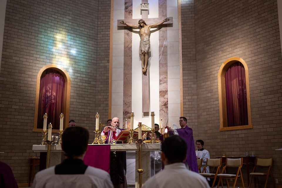 Bishop conducts mass in a church with a crucifix and stained glass light. Clergy in purple vestments, altar with candles and gold details.