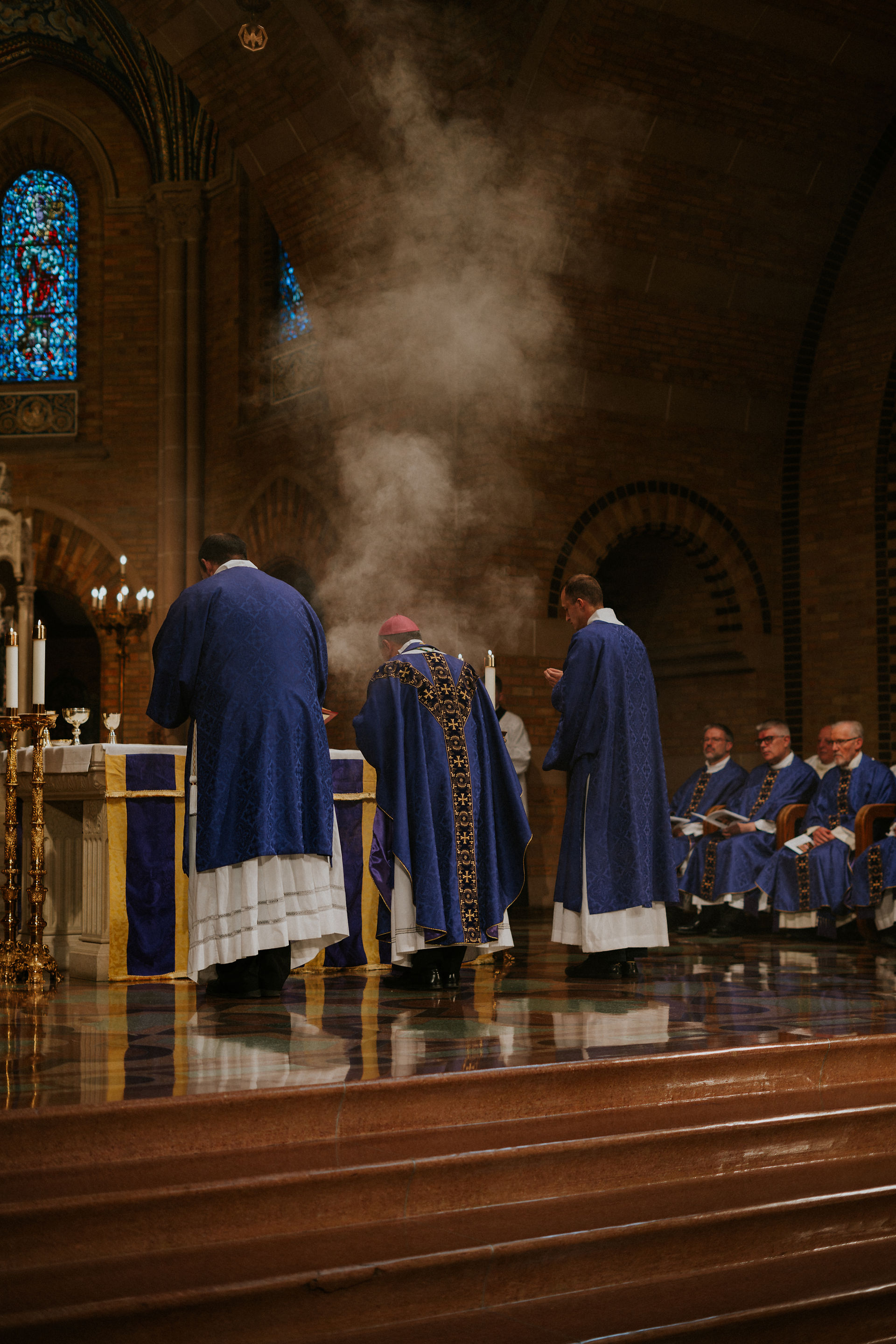 Priests in purple vestments conduct a ceremony at an altar, incense smoke rising. Stained glass and candlelight create a solemn atmosphere.