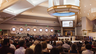 A congregation inside a church, seated and standing, with a screen displaying "Twentieth Sunday in Ordinary Time." Warm lighting and organ pipes seen.